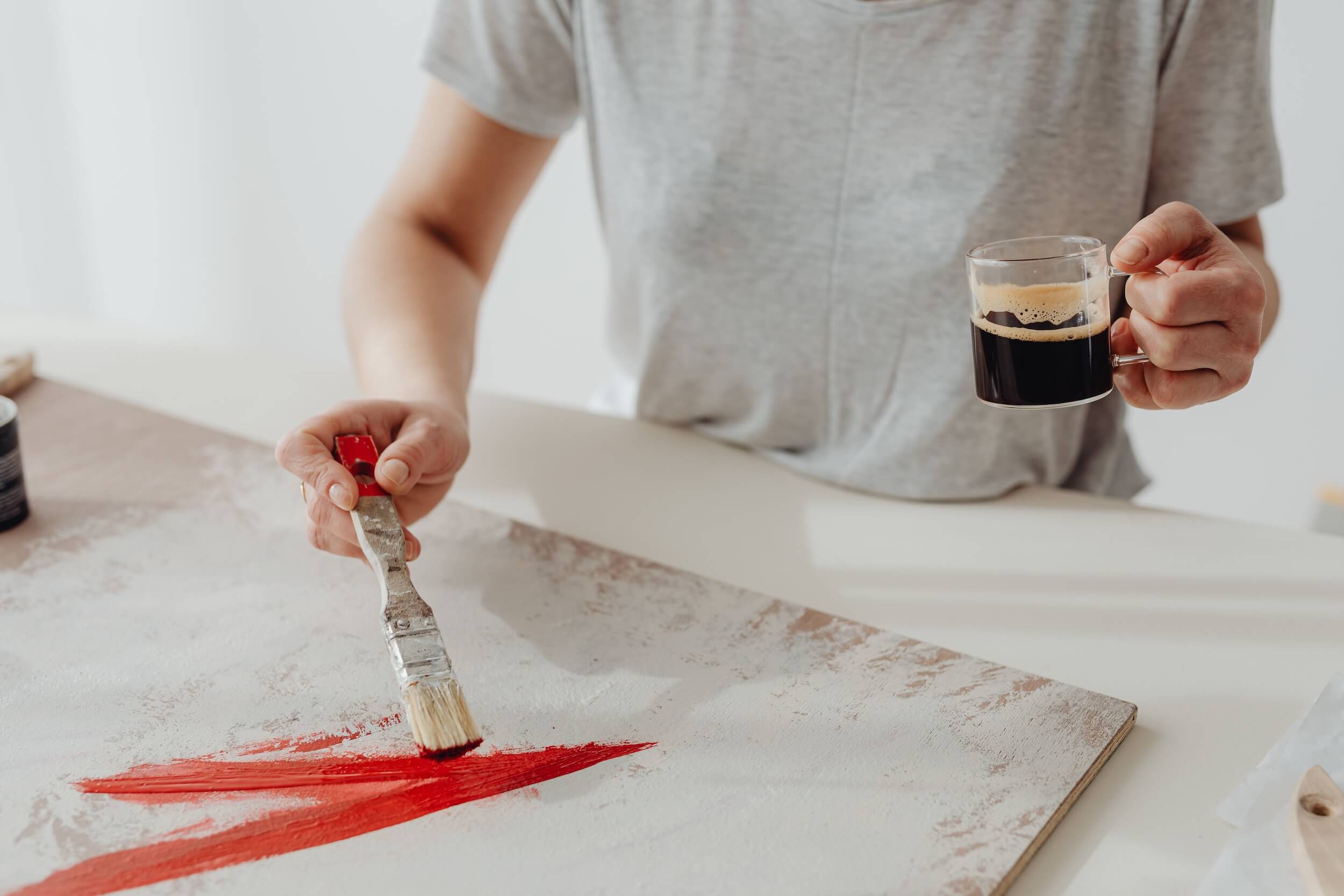 Frau streicht eine Möbelplatte mit Farbe und hält in der anderen Hand eine Tasse Kaffee